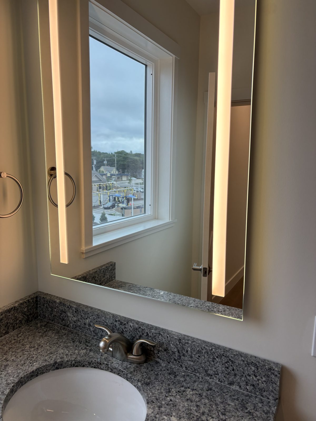 A backlit LED vanity mirror above a granite bathroom counter in a GALE residence, reflecting a window and the bathroom door