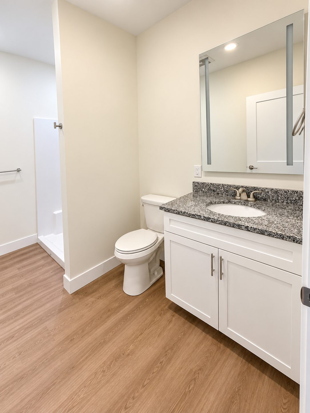 A GALE bathroom with a walk-in shower, granite vanity with white shaker cabinet, and a backlit mirror, with wood-look plank floors