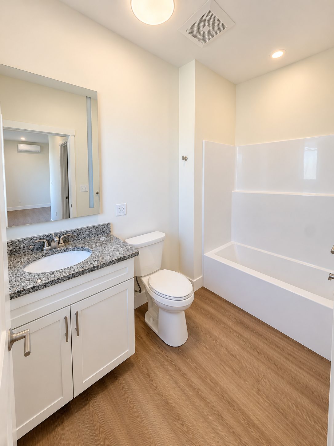 A GALE bathroom with a granite vanity, white shaker cabinet, a backlit mirror, and a white tub and shower surround, with wood-look plank floors