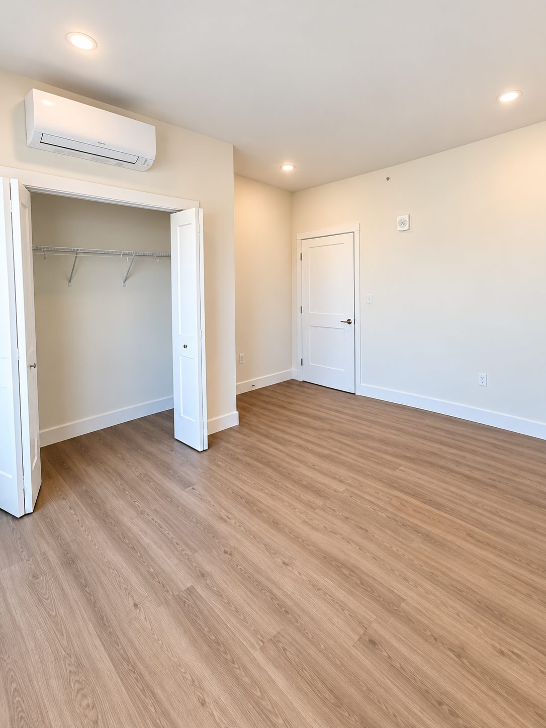A bedroom in a GALE residence with a wall-mounted ductless mini-split above a bifold double closet, recessed lighting, and wood-look plank floors